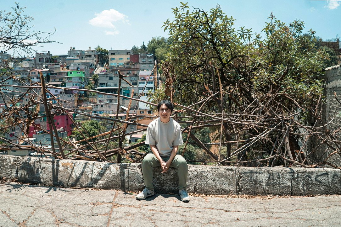 Armando Maravilla sentado sobre un muro de concreto en una barranca del poniente de la Ciudad de México, con ramas secas detrás y casas en la ladera